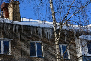 Big beautiful icicles hang from the snow-covered roof in front of the windows of the house. Snow on the roof. Frosty sunny day