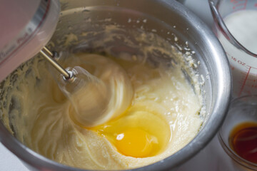 Making of a cake: Adding an egg and beating batter.  Mixer beaters showing motion blur.  Milk and vanilla in background.