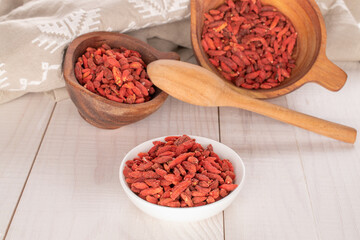 Dried goji berries in a white saucer and in two wooden cups with a wooden spoon on a wooden table, macro.