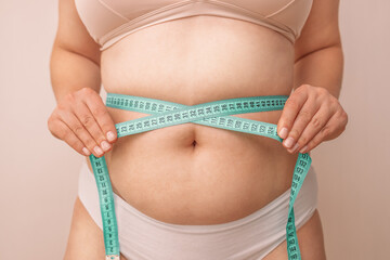 Fat belly. Fat caucasian girl measuring waist with green measuring tape on a beige background,...