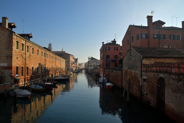 Naklejka na ściany i meble Ein Wasserkanal zieht sich vom Meer in den Venezianischen Stadtteil Castello. Boote liegen am Uferweg und bei Blauem Himmel ist noch der Frühnebel zu erkennen der sich auflöst.