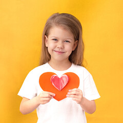 Child kid holding red heart to St Valentine day handmade from paper, on yellow background waist up caucasian little girl of 5 years in white t-shirt