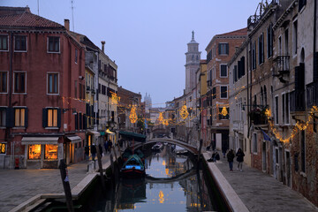 Leute laufen an Wasserkanal in Venedig entlang und weihnachtliche Beleuchtung spiegelt sich im...