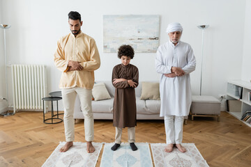 Fototapeta premium Muslim kid crossing arms while praying near interracial family at home.
