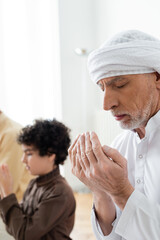 Mature man praying with closed eyes near blurred arabian grandson at home.