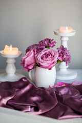 Fototapeta na wymiar Bouquet of purple roses in a white vase against a background of purple fabric and candles, still life