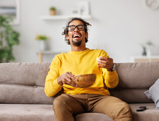 Young overjoed african american man with popcorn laughing out of loud, watching comedy movie at home