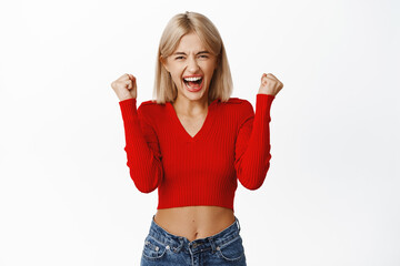 Image of enthusiastic lucky blond girl winning, shouting with joy and fist pump, feeling powerful...