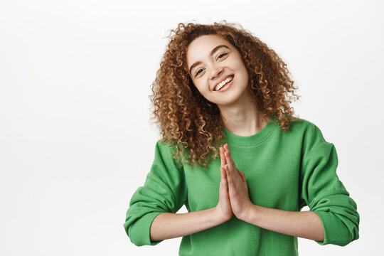 Thank you. Smiling beautiful curly girl, holding hands clasped and looking pleased, being grateful, thankful, showing namaste gesture, standing over white background