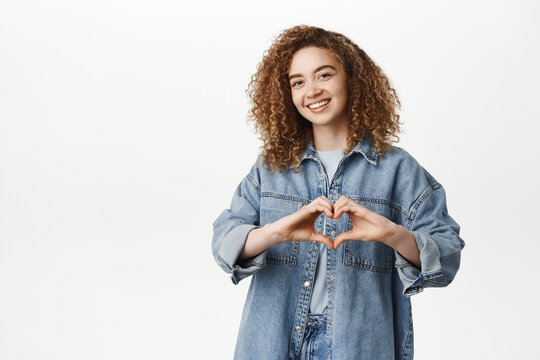 Portrait of smiling curly girl, stylish girlfriend shows heart, love sign and pucker lips, kissing gesture, standing in denim clothes over white background