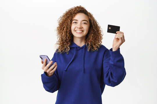 Happy young woman shows credit card and smartphone, smiles at camera, concept of mobile banking, online shopping and apps, standing over white background