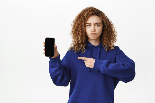 Angry young woman frowning, pointing at smartphone screen, mobile phone app, complaining at smth, standing in blue hoodie over white background