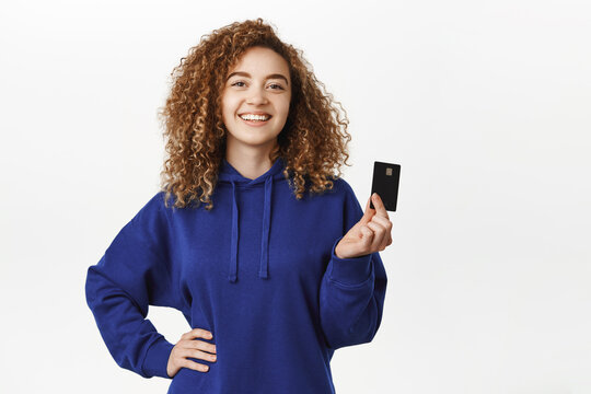 Portrait of amazed curly girl shows credit card and smiles, demonstrates new bank feature, recommending, standing against white background