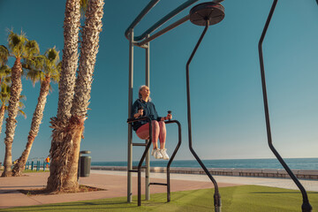 Adult female holding on uneven bars and raising her knees up on the sports field by the seashore