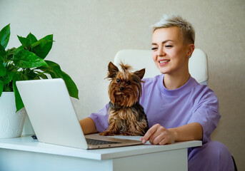 a young woman, blonde with short hair,sits a lady in purple pajamas,near a laptop, and holds a small dog in her arms