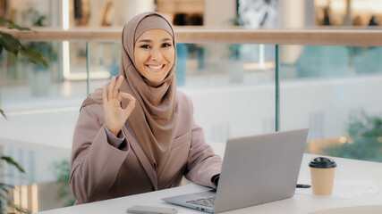 Confident muslim businesswoman freelancer in hijab working typing on laptop chatting makes purchases in online store girl student studying remotely shows OK gesture approves new computer application