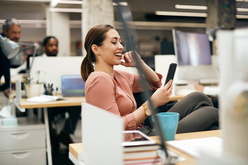 Young businesswoman laughs while reading text message on cell phone in office.
