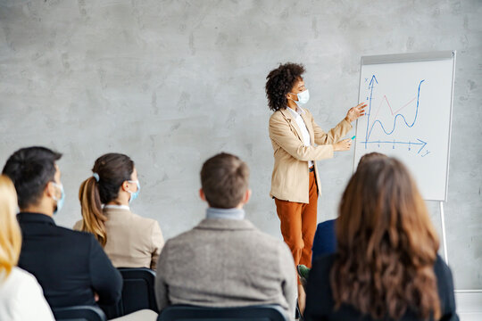 Businesswoman seminar and presentation. An African American corporate businesswoman with a face mask standing next to a chart board and pointing at statistics in front of the people.