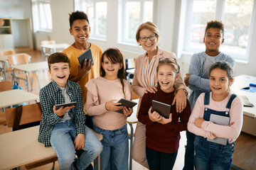 Group of happy elementary students with touchpads and their teacher in the classroom at school. - obrazy, fototapety, plakaty