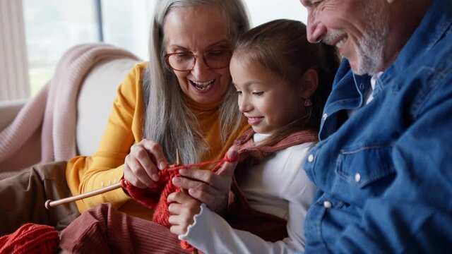 Little girl sitting on sofa with her grandparents and learning to knit indoors at home.