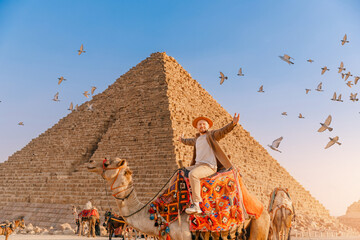 Tourist man with hat riding on camel background pyramid of Egyptian Giza, sunset Cairo, Egypt - obrazy, fototapety, plakaty