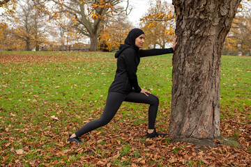 Woman in black sports clothing and hijab stretching at tree trunk in park