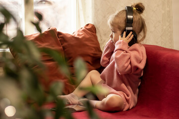 a little cute girl with headphones is sitting on the couch and listening to music on a mobile tablet. the child holds headphones with his hands. a girl  looks out the window