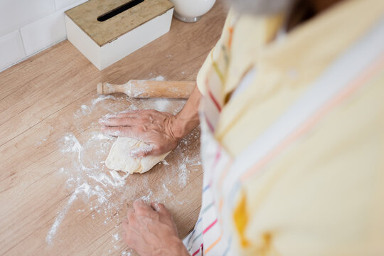 High angle view of mature man cooking dough near flour and rolling pin on worktop in kitchen.