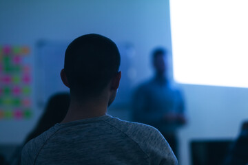 Back view of students during class during lecture. Professor next to a projected screen on the wall of a modern classroom..
