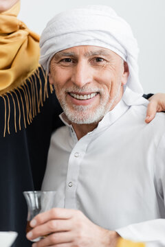 cheerful muslim man in turban holding cup of tea and looking at camera near wife at home.