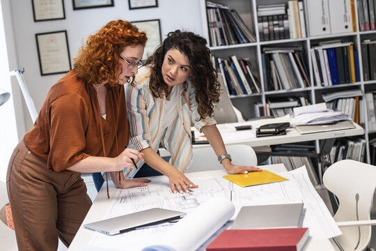 Two female coworkers are trying to solve a problem of a new project.They were standing by the desk and looking at the blueprint.Working day at the office.	