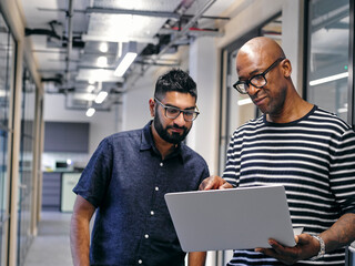 Two men in office hallway, looking at laptop