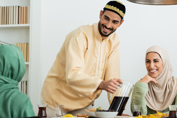 happy muslim man in skullcap pouring tea near happy woman in hijab.