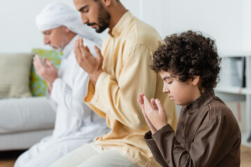 Side view of curly muslim kid praying near interracial parents at home.