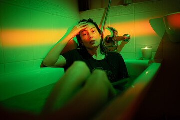 Young depressed asian woman in neon light sitting in bathtub filled with water, suffering from...