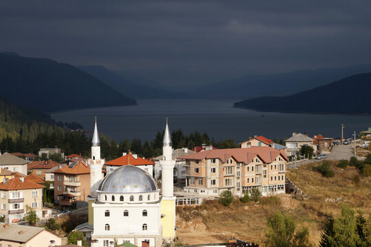 a small town with a mosque in sunlight against a stormy sky, mountains and a lake. Lake Dospat Bulgaria