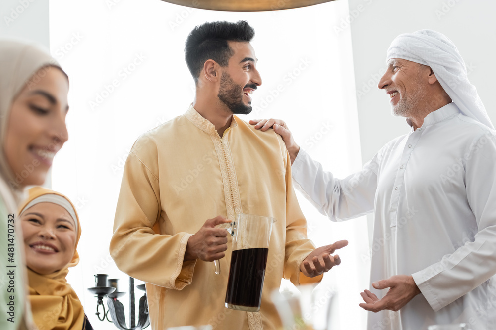 Wall mural excited arabian man holding jug of tea while talking to father near interracial family.