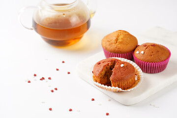 Several homemade white and pink muffins on marble board on white surface with heart-shaped red and white sugar decoration and tea-pot