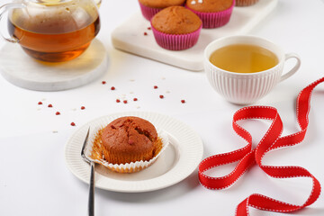 Several homemade white and pink muffins on marble board on white surface, red ribbon, heart-shaped red and white sugar decoration and tea-pot