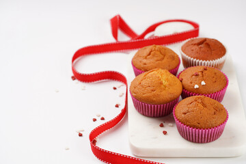 Several homemade white and pink muffins on marble board on white surface, red ribbon, heart-shaped red and white sugar decoration and tea-pot