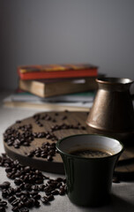 on a linen tablecloth a green cup of coffee, a turk for making coffee on a wooden stand, grains scattered next to it and books on in the background