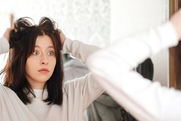 A young woman stands in front of a mirror and looks in surprise at the condition of her hair, seasonal problem of hair loss, oily, dry, unkempt hair