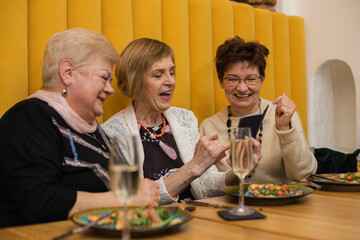 Laughing elderly dark and fair-haired women looking at phone, having pizza on plates and champagne in glass, dicussing photo while celebrating their meeting or holiday in modern cafe with orange walls