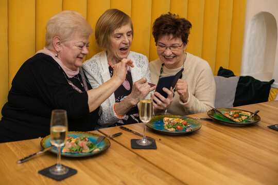 Smiling elderly dark and fair-haired women looking at phone, having pizza on plates, dicussing photos while celebrating their meeting or holiday in modern cafe with orange walls