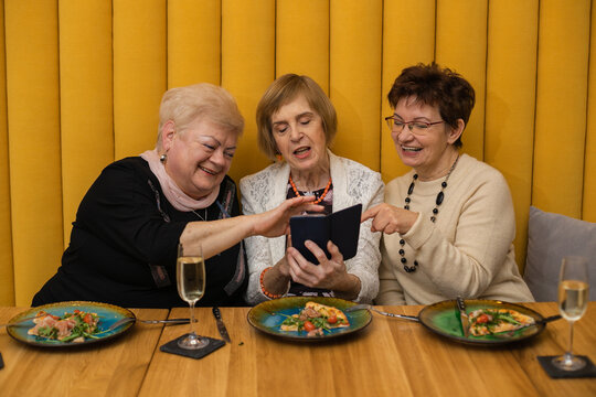 Relaxing elderly women dressed in white, beige and black looking at phone, having pizza on plates, dicussing photos while celebrating their meeting or holiday in modern cafe with orange walls