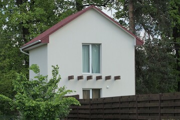facade of a white private concrete house with windows under a red tiled roof behind a brown wooden fence among green trees