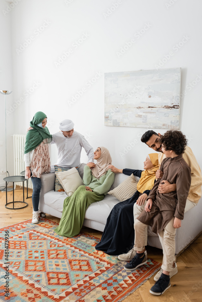 Wall mural young muslim man hugging arabian son near cheerful multicultural family in living room.