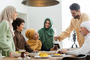 Arabian man pouring tea near food and smiling interracial family at home.