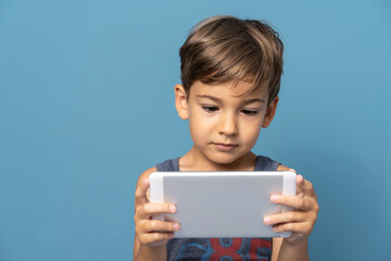 Front view of small caucasian boy four years old standing in front of blue background studio shot standing and using digital tablet looking to the digital screen happy serious