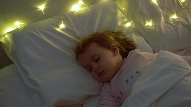 Cute Chuby Little Toddler Baby Girl 1-2 Years Old Sleeping In Pink Pajama On White Bed In Dark Bedroom. Child Have Relax Rest Time At Night Garland on Pillow. Childhood, Christmas, Parenthhood Concept